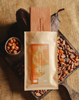 Tablette de chocolat au lait au noisettes de lot et garonne Origines Chocolate Makers, emballée dans un sachet kraft avec une étiquette marron indiquant 38 % de cacao de Sâo Tomé entourée de fèves de cacao, de cabosses et d'un bol contenant des noisettes sur un fond en toile de jute.