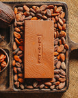 Tablette de chocolat au lait au noisettes de lot et garonne Origines Chocolate Makers,38 % de cacao de São Tomé entourée de fèves de cacao, de cabosses et d'un bol contenant des noisettes sur un fond en toile de jute.