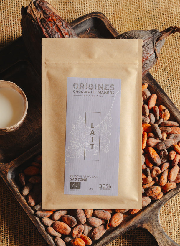 Tablette de chocolat au lait Origines Chocolate Makers, emballée dans un sachet kraft avec une étiquette violette indiquant 38 % de cacao provenant de São Tomé, entourée de fèves de cacao, de cabosses et d'un bol contenant du lait sur un fond en toile de jute.