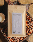 Tablette de chocolat au lait Origines Chocolate Makers, emballée dans un sachet kraft avec une étiquette violette indiquant 38 % de cacao provenant de São Tomé, entourée de fèves de cacao, de cabosses et d'un bol contenant du lait sur un fond en toile de jute.