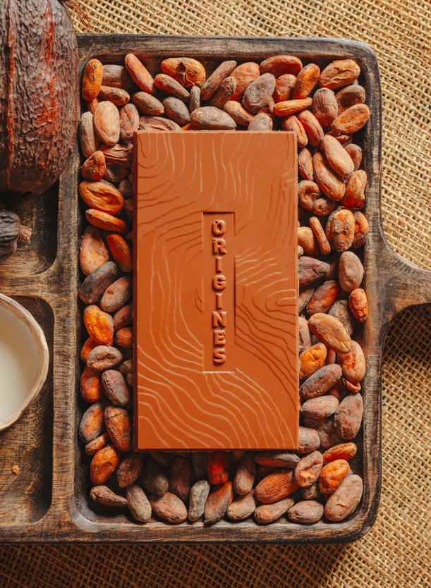 Tablette de chocolat au lait Origines Chocolate Makers 55 % de cacao provenant de Madagascar, entourée de fèves de cacao, de cabosses et d'un bol contenant du lait sur un fond en toile de jute.