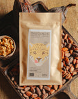 Tablette de chocolat au lait au céréales croustillantes Origines Chocolate Makers, emballée dans un sachet kraft avec une étiquette représentant un tigre indiquant 38 % de cacao provenant de Sao Tome, entourée de fèves de cacao, de cabosses et d'un bol contenant des céréales sur un fond en toile de jute.