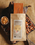 Tablette de chocolat au lait au céréales croustillantes Origines Chocolate Makers, emballée dans un sachet kraft avec une étiquette représentant un tigre indiquant 38 % de cacao provenant de Sao Tome, entourée de fèves de cacao, de cabosses et d'un bol contenant des céréales sur un fond en toile de jute.