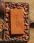 Tablette de chocolat au lait au céréales croustillantes Origines Chocolate Makers, 38 % de cacao provenant de Sao Tome, entourée de fèves de cacao, de cabosses et d'un bol contenant des céréales sur un fond en toile de jute.