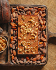 Dos d'une tablette de chocolat au lait au céréales croustillantes Origines Chocolate Makers, 38 % de cacao provenant de Sao Tome, entourée de fèves de cacao, de cabosses et d'un bol contenant des céréales sur un fond en toile de jute.