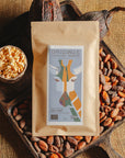 Tablette de chocolat au lait au riz soufflé Origines Chocolate Makers, emballée dans un sachet kraft avec une étiquette représentant une girafe indiquant 38 % de cacao provenant de Sao Tome, entourée de fèves de cacao, de cabosses et d'un bol contenant du riz soufflé sur un fond en toile de jute.