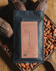 Tablette de chocolat noir Origines Chocolate Makers, emballée dans un sachet noir avec une étiquette rouge indiquant 70 % de cacao provenant de Sao Tome, entourée de fèves de cacao et de cabosses sur un fond en toile de jute.