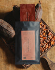 Tablette de chocolat noir Origines Chocolate Makers, emballée dans un sachet noir avec une étiquette rouge indiquant 70 % de cacao provenant de Sao Tome, entourée de fèves de cacao et de cabosses sur un fond en toile de jute.