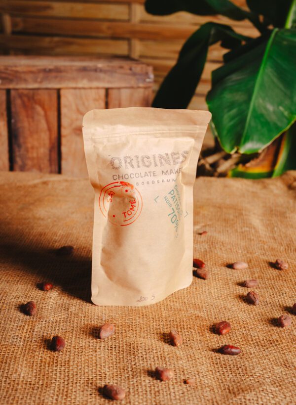 Un sachet de chocolat pâtissier en papier kraft avec l'inscription "Origines Chocolate Maker Bordeaux" sur une table en toile de jute. Autour du sachet, on peut voir des fèves de cacao éparpillées. L'arrière-plan présente une caisse en bois et une plante verte avec de grandes feuilles, créant une ambiance naturelle et artisanale.