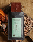 Tablette de chocolat noir Origines Chocolate Makers, emballée dans un sachet noir avec une étiquette bleue indiquant 70 % de cacao provenant de la coopérative Cabruca à Bahia, Brésil, entourée de fèves de cacao et de cabosses sur un fond en toile de jute.