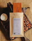 Tablette de chocolat au lait Origines Chocolate Makers, emballée dans un sachet kraft avec une étiquette violette indiquant 38 % de cacao provenant de São Tomé, entourée de fèves de cacao, de cabosses et d'un bol contenant du lait sur un fond en toile de jute.