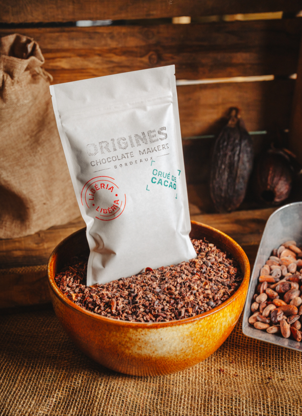 Un sachet de grué de cacao en papier blanc avec l'inscription "Origines Chocolate Maker Bordeaux" posé dans un bol de grué de cacao et sur une table en toile de jute. L'arrière-plan présente  une ambiance naturelle et artisanale avec des cabosses de cacao et un sac en jute.