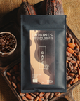 Tablette de chocolat noir au café Origines Chocolate Makers, emballée dans un sachet noir avec une étiquette marron indiquant 70 % de cacao provenant de Colombie, entourée de fèves de cacao, de cabosses et d'un bol contenant du café sur un fond en toile de jute.