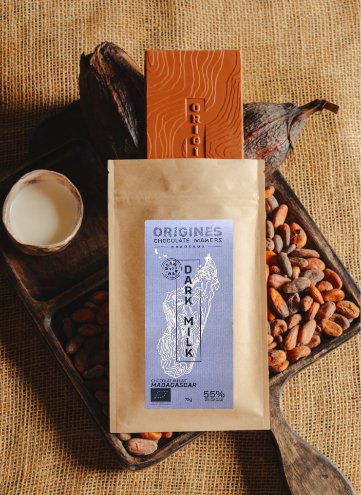 Tablette de chocolat au lait Origines Chocolate Makers, emballée dans un sachet kraft avec une étiquette violette indiquant 55 % de cacao provenant de Madagascar, entourée de fèves de cacao, de cabosses et d'un bol contenant du lait sur un fond en toile de jute.