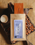 Tablette de chocolat au lait Origines Chocolate Makers, emballée dans un sachet kraft avec une étiquette violette indiquant 55 % de cacao provenant de Madagascar, entourée de fèves de cacao, de cabosses et d'un bol contenant du lait sur un fond en toile de jute.