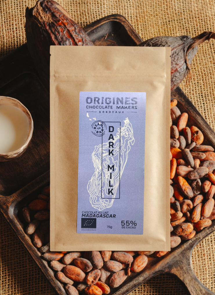 Tablette de chocolat au lait Origines Chocolate Makers, emballée dans un sachet kraft avec une étiquette violette indiquant 55 % de cacao provenant de Madagascar, entourée de fèves de cacao, de cabosses et d'un bol contenant du lait sur un fond en toile de jute.