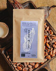 Tablette de chocolat au lait Origines Chocolate Makers, emballée dans un sachet kraft avec une étiquette violette indiquant 55 % de cacao provenant de Madagascar, entourée de fèves de cacao, de cabosses et d'un bol contenant du lait sur un fond en toile de jute.