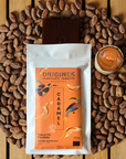 Tablette de chocolat noir Origines Chocolate Makers au caramel, emballée dans un sachet orange orné de toucans colorés, entourée de fèves de cacao et d'un petit bol de caramel sur une surface en bois.