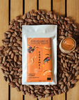 Tablette de chocolat noir Origines Chocolate Makers au caramel, emballée dans un sachet orange orné de toucans colorés, entourée de fèves de cacao et d'un petit bol de caramel sur une surface en bois.