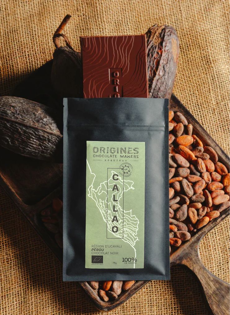 Tablette de chocolat noir Origines Chocolate Makers, emballée dans un sachet noir avec une étiquette verte indiquant 100 % de cacao provenant du Pérou, entourée de fèves de cacao et de cabosses sur un fond en toile de jute.