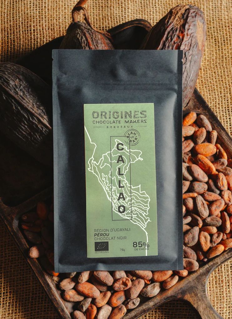 Tablette de chocolat noir Origines Chocolate Makers, emballée dans un sachet noir avec une étiquette verte indiquant 85 % de cacao provenant du Pérou, entourée de fèves de cacao et de cabosses sur un fond en toile de jute.
