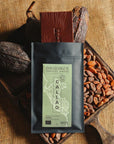 Tablette de chocolat noir Origines Chocolate Makers, emballée dans un sachet noir avec une étiquette verte indiquant 100 % de cacao provenant du Pérou, entourée de fèves de cacao et de cabosses sur un fond en toile de jute.