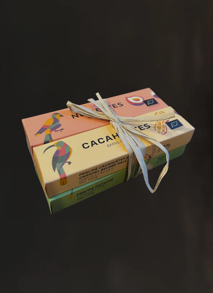 Un coffret de 4 boites de barres chocolatées à la cacahuètes, à la noisette, à la pistache et au caramel. Il y a des oiseaux dessinés sur les boites et elles sont entourées avec un ruban en paille.