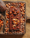 Tablette de chocolat noir au amandes de Valence Origines Chocolate Makers 70 % de cacao provenant du Pérou, entourée de fèves de cacao, de cabosses et d'un bol contenant des amandes sur un fond en toile de jute.