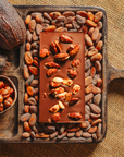 Tablette de chocolat au lait au amandes de Valence Origines Chocolate Makers,38 % de cacao de São Tomé entourée de fèves de cacao, de cabosses et d'un bol contenant des amandes sur un fond en toile de jute.