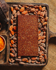 Tablette de chocolat noir au piment d'Espelette Origines Chocolate Makers 70 % de cacao provenant du Pérou, entourée de fèves de cacao, de cabosses et d'un bol contenant du piment d'Espelette sur un fond en toile de jute.