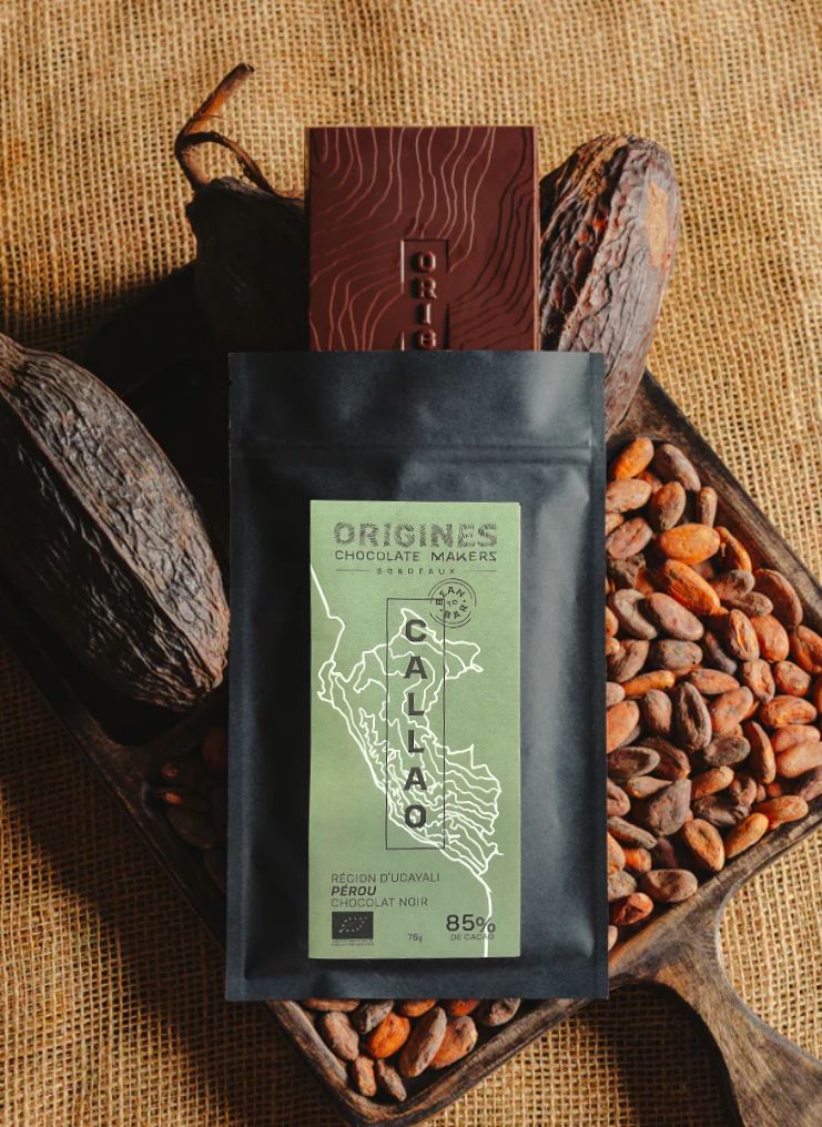 Tablette de chocolat noir Origines Chocolate Makers, emballée dans un sachet noir avec une étiquette verte indiquant 85% de cacao provenant du Pérou, entourée de fèves de cacao et de cabosses sur un fond en toile de jute.