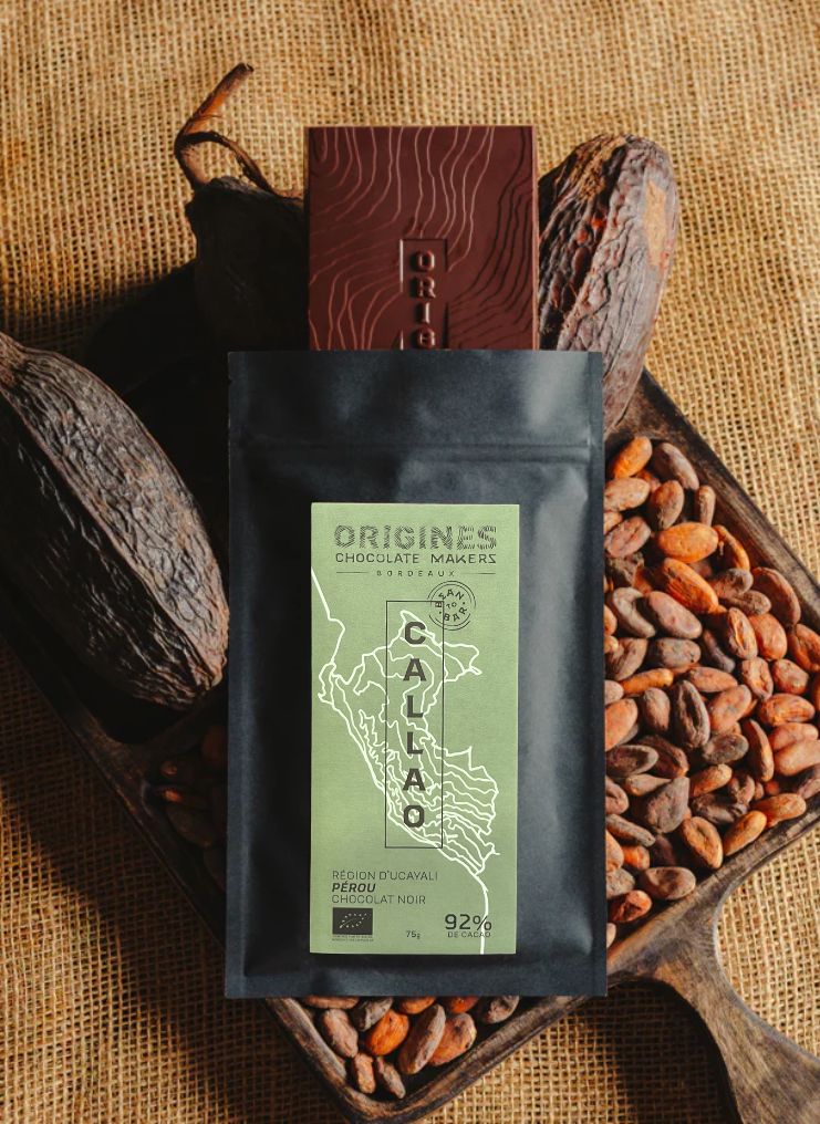 Tablette de chocolat noir Origines Chocolate Makers, emballée dans un sachet noir avec une étiquette verte indiquant 92 % de cacao provenant du Pérou, entourée de fèves de cacao et de cabosses sur un fond en toile de jute. Nous apercevons la tablette sortir de son emballage.
