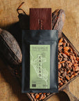 Tablette de chocolat noir Origines Chocolate Makers, emballée dans un sachet noir avec une étiquette verte indiquant 92 % de cacao provenant du Pérou, entourée de fèves de cacao et de cabosses sur un fond en toile de jute. Nous apercevons la tablette sortir de son emballage.