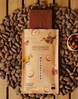 Tablette de chocolat noir Origines Chocolate Makers à la noisette, emballée dans un sachet orange orné de toucans colorés, entourée de fèves de cacao et d'un petit bol de noisettes sur une surface en bois.