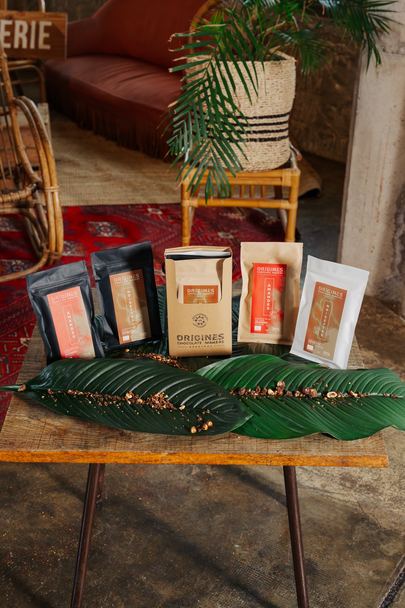 Produits Origines Chocolate Makers, comprenant plusieurs paquets de chocolat posés sur des feuilles vertes, sur une table en bois dans un cadre chaleureux avec un tapis rouge et des plantes