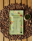 Tablette de chocolat noir Origines Chocolate Makers à la pistache, emballée dans un sachet vert orné de toucans colorés, entourée de fèves de cacao et d'un petit bol de pistaches sur une surface en bois.