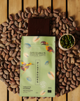 Tablette de chocolat noir Origines Chocolate Makers à la pistache, emballée dans un sachet vert orné de toucans colorés, entourée de fèves de cacao et d'un petit bol de pistaches sur une surface en bois.