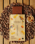 Tablette de chocolat noir Origines Chocolate Makers à la cacahuète, emballée dans un sachet jaune orné de toucans colorés, entourée de fèves de cacao et d'un petit bol de cacahuètes sur une surface en bois.
