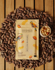 Tablette de chocolat noir Origines Chocolate Makers à la cacahuète, emballée dans un sachet jaune orné de toucans colorés, entourée de fèves de cacao et d'un petit bol de cacahuètes sur une surface en bois.