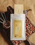 Tablette de chocolat blanc à la vanille Origines Chocolate Makers, emballée dans un sachet blanc avec une étiquette jaune indiquant 35% de cacao provenant de Madagascar, entourée de fèves de cacao, de gousses de vanille et de cabosses sur un fond en toile de jute.