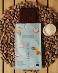 Tablette de chocolat noir Origines Chocolate Makers à la noix de coco, emballée dans un sachet bleuorné de toucans colorés, entourée de fèves de cacao et d'un petit bol de noix de coco sur une surface en bois.