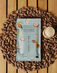 Tablette de chocolat noir Origines Chocolate Makers à la noix de coco, emballée dans un sachet bleuorné de toucans colorés, entourée de fèves de cacao et d'un petit bol de noix de coco sur une surface en bois.
