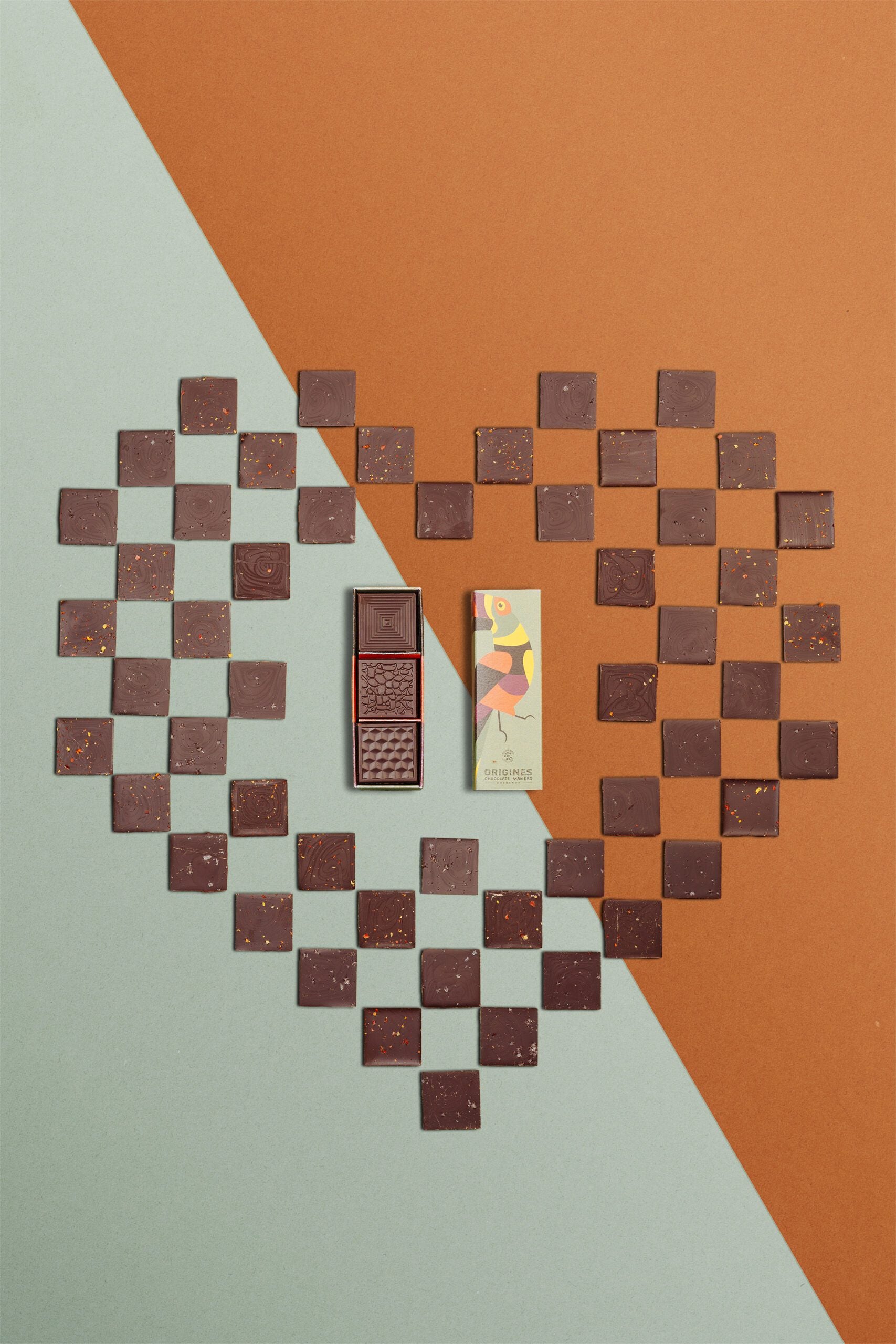 Une boîte rectangulaire de chocolats fins avec un toucan sur l'emballage est ouverte, révélant trois carrés de chocolat aux motifs variés, posée sur un cœur fait avec les carrés de chocolat et un fond géométrique dans les tons orange et vert.