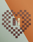 Une boîte rectangulaire de chocolats fins avec un toucan sur l'emballage est ouverte, révélant trois carrés de chocolat aux motifs variés, posée sur un cœur fait avec les carrés de chocolat et un fond géométrique dans les tons orange et vert.