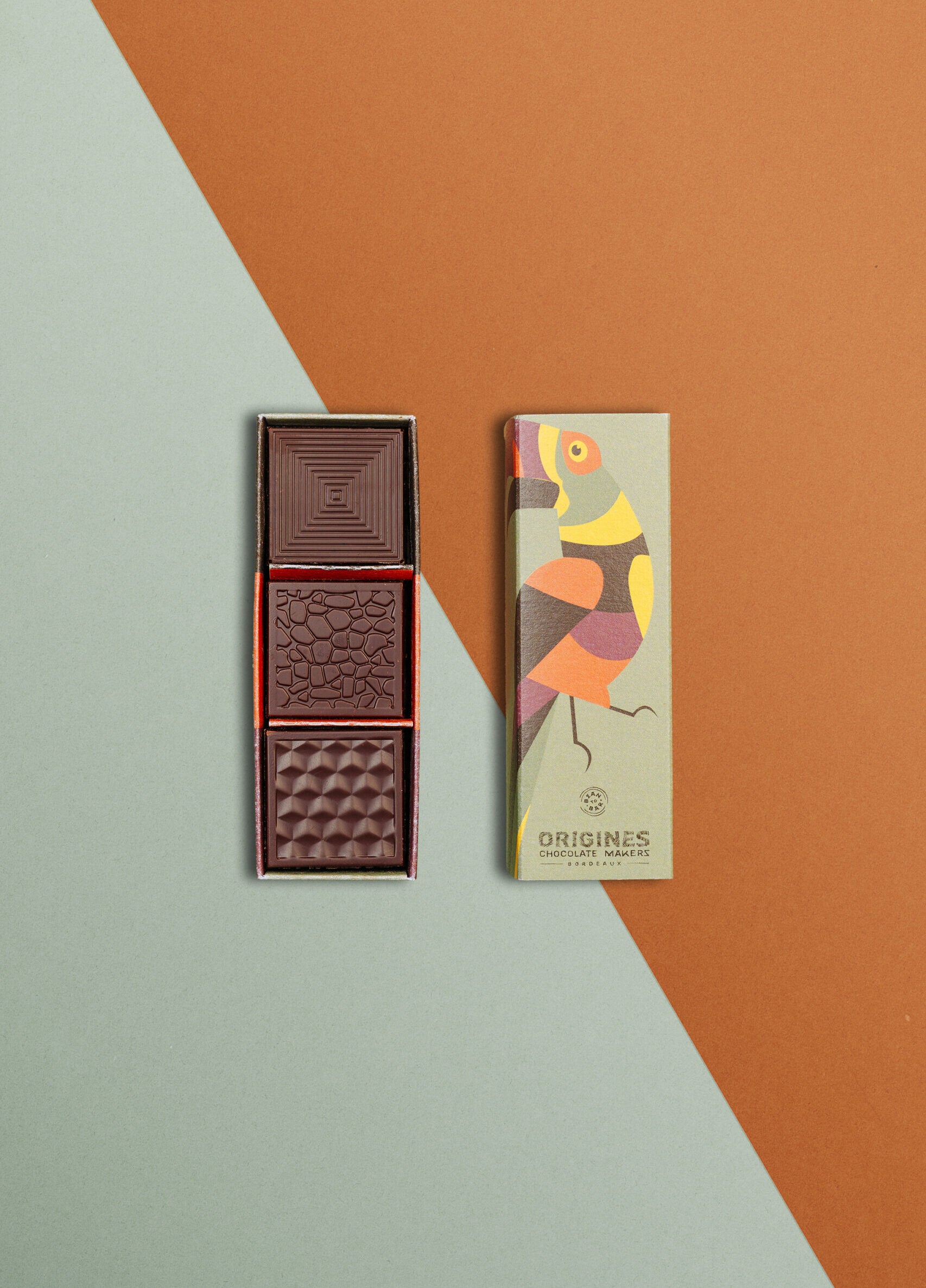 Une boîte rectangulaire de chocolats fins avec un toucan sur l'emballage est ouverte, révélant trois carrés de chocolat aux motifs variés, posée sur un fond géométrique dans les tons orange et vert.