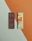 Une boîte rectangulaire de chocolats fins avec un toucan sur l'emballage est ouverte, révélant trois carrés de chocolat aux motifs variés, posée sur un fond géométrique dans les tons orange et vert.