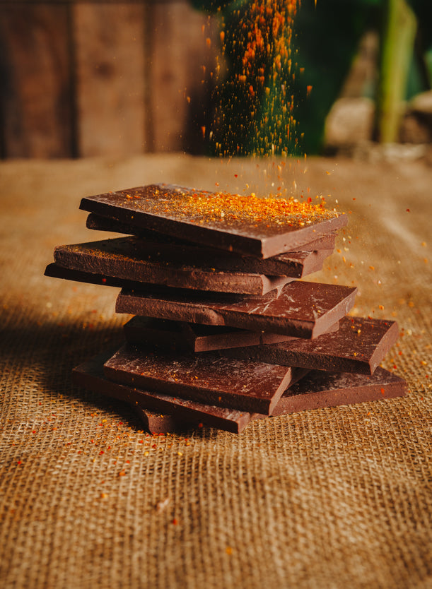 Morceaux de chocolat noir aux piment d'Espelette empilés sur une toile de jute, avec du piment qui tombe sur le chocolat, en arrière-plan naturel.