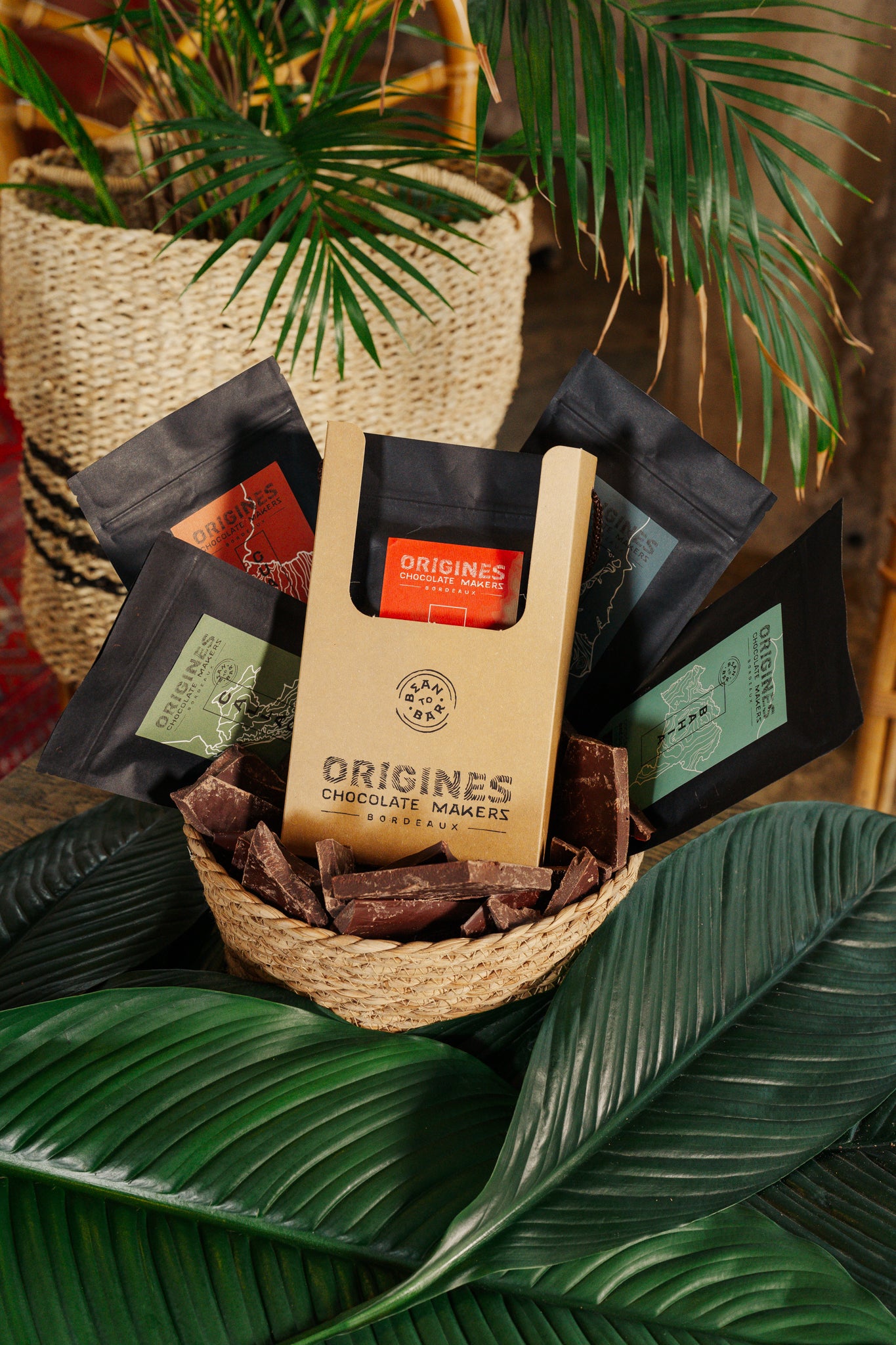 Produits Origines Chocolate Makers, comprenant plusieurs paquets de chocolat posés sur des feuilles vertes, sur une table en bois dans un cadre chaleureux.