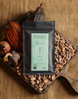 Tablette de chocolat noir Origines Chocolate Makers, emballée dans un sachet noir avec une étiquette verte indiquant 70 % de cacao provenant de la coopérative Cabruca à Bahia, Brésil, entourée de fèves de cacao et de cabosses sur un fond en toile de jute.