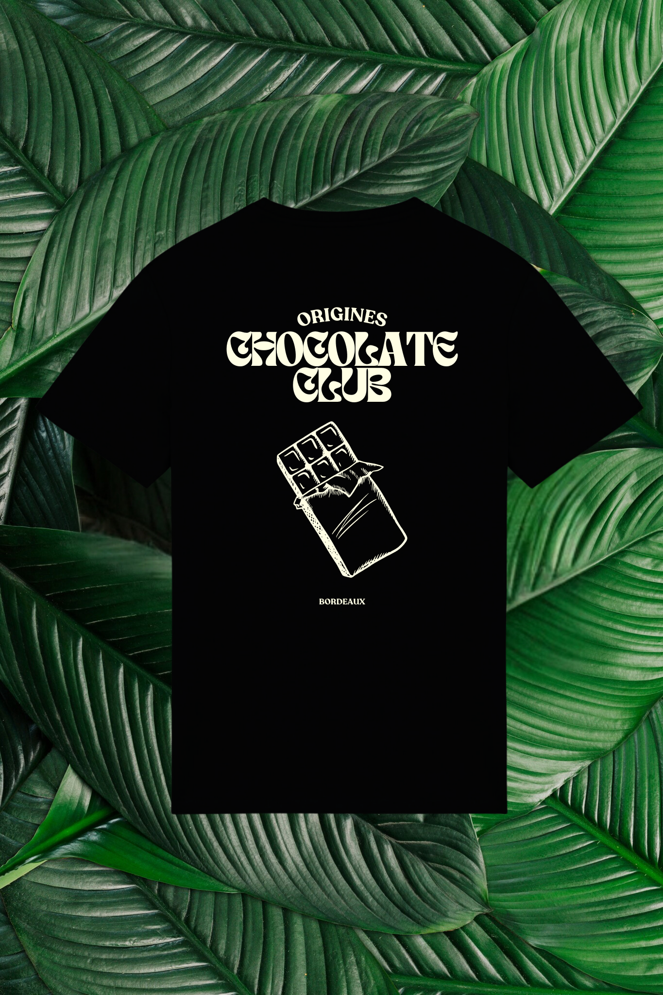 Un t-shirt noir avec un design au dos représentant une tablette de chocolat illustrée de façon minimaliste. Le texte 'Origines Chocolate Club' est inscrit en haut en lettres stylisées, suivi de l'inscription 'Bordeaux' en bas. Le t-shirt est posé sur un fond de grandes feuilles vertes naturelles.